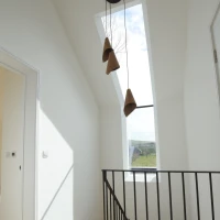 Pasillo iluminado con pared blanca y escalera de madera. Ventana de techo que permite la entrada de luz natural. Tres lámparas colgantes de forma cónica.