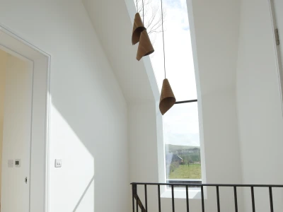 Un couloir lumineux avec un plafond en pente et une grande fenêtre ouverte sur le ciel. Trois suspensions en forme de cône sont accrochées au plafond. Un escalier en bois est visible à gauche, avec une balustrade noire minimaliste.