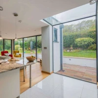 A modern open-plan living space featuring a kitchen with granite countertops, a dining area with a vase of flowers, and large glass windows showcasing a green garden. Natural light floods the room, highlighting contemporary furniture and decor.