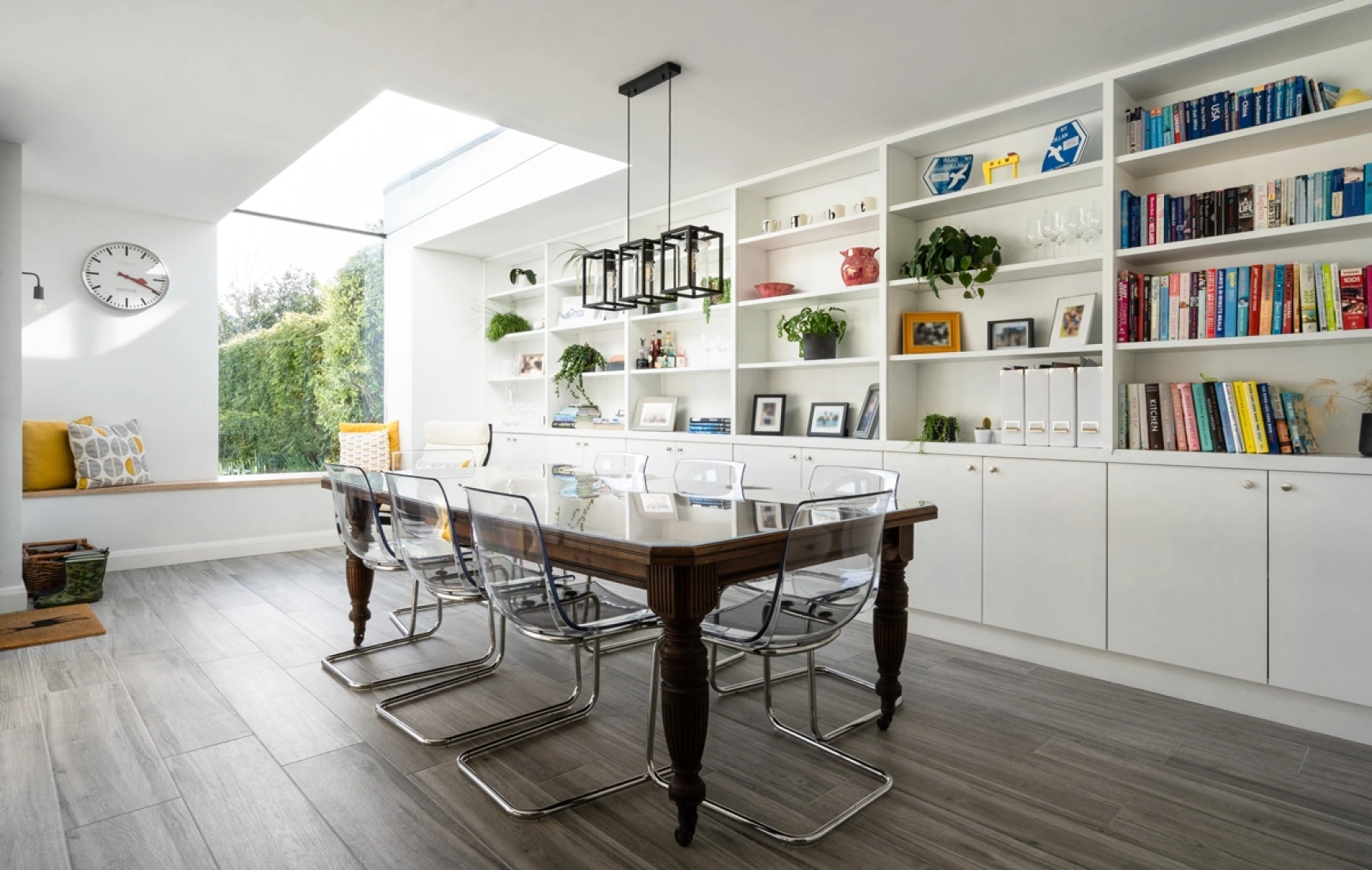 Helle, moderne Küche mit einem langen Holztisch und transparenten Stühlen. An einer Wand sind Regale mit Pflanzen und Büchern. Ein Fenster im Dach bringt zusätzliches Licht hinein.