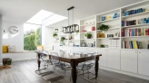 Comedor elegante con mesa de madera y sillas transparentes. Estanterías blancas llenas de libros y decoraciones. Ventana grande que ilumina el espacio. Detalles como plantas y un rinconcito con almohadas.