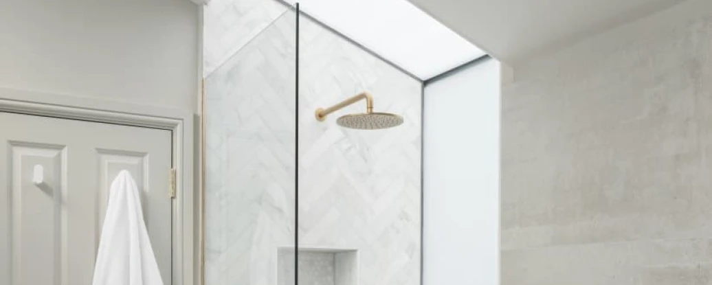 A modern shower with a glass enclosure features a gold showerhead and fixtures. The walls are tiled with white marble in a herringbone pattern, and a window above provides natural light. A folded white towel hangs on the door outside the shower.