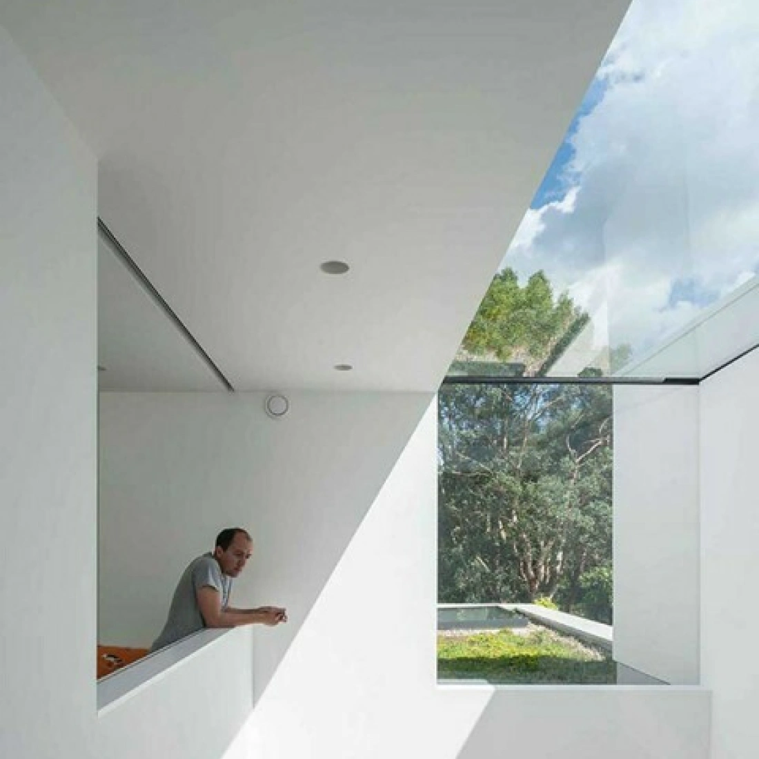 Un homme se penche sur une balustrade dans un espace intérieur moderne, avec de grandes fenêtres laissant entrer la lumière naturelle et une vue sur la végétation extérieure.