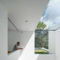 Un homme se penche sur une balustrade dans un espace intérieur moderne, avec de grandes fenêtres laissant entrer la lumière naturelle et une vue sur la végétation extérieure.