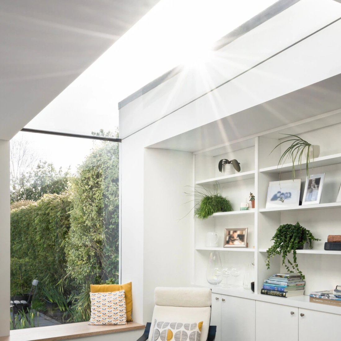 Espacio luminoso con un sillón blanco y negro, estanterías con decoraciones y plantas. Ventana amplia que permite la entrada de luz solar y vistas al jardín exterior. Suelo de madera clara.