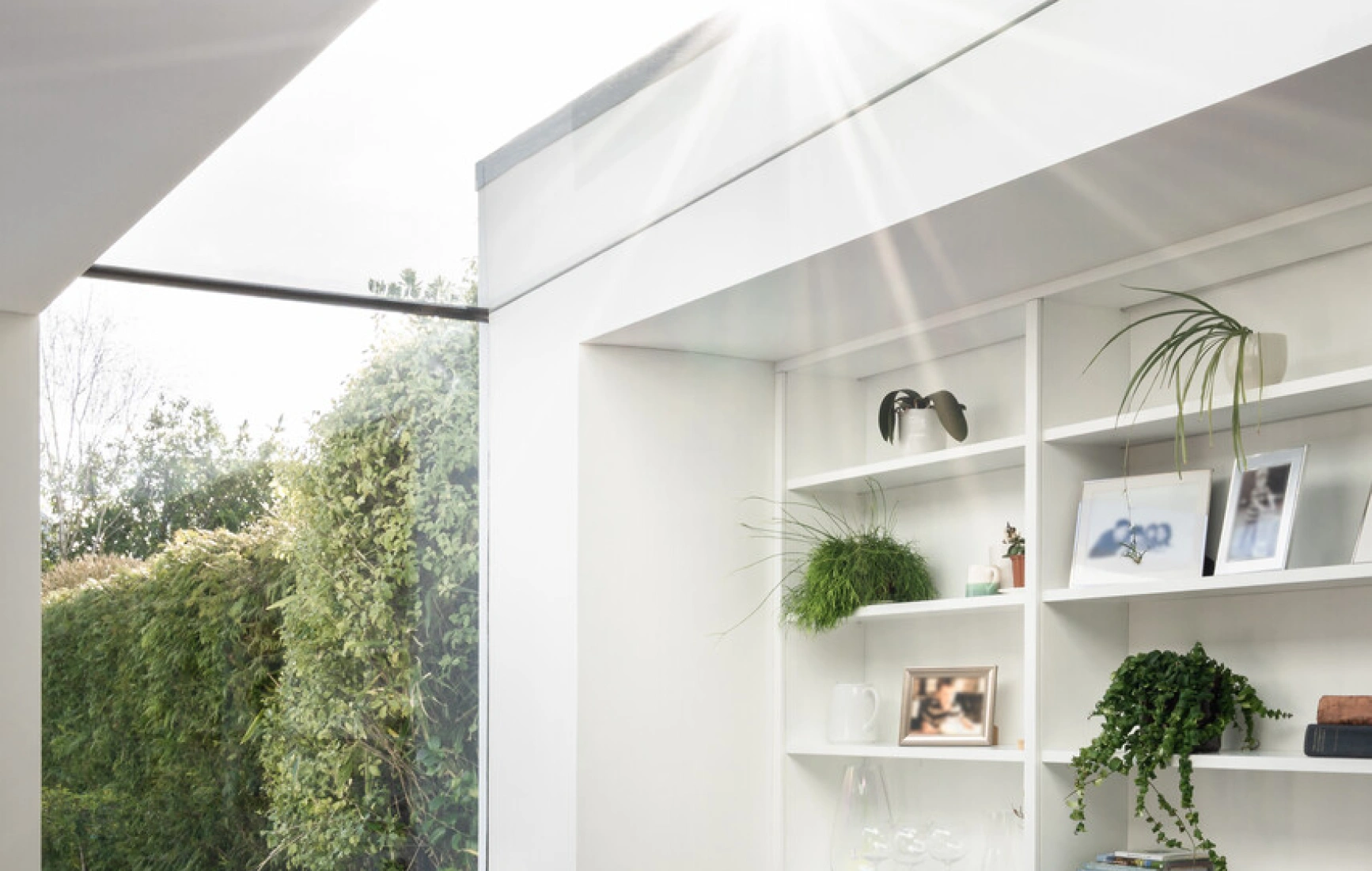 Un coin lumineux d'une pièce moderne avec une chaise confortable, des étagères blanches contenant des plantes et des objets décoratifs. De grandes fenêtres laissent entrer la lumière naturelle, offrant une vue intérieure-extérieur sur un jardin verdoyant.