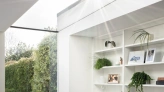 Espacio luminoso con un sillón blanco y negro, estanterías con decoraciones y plantas. Ventana amplia que permite la entrada de luz solar y vistas al jardín exterior. Suelo de madera clara.