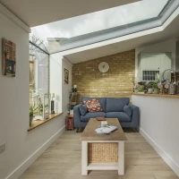 A cozy living space featuring a blue sofa, a wooden coffee table, and a brick wall. Natural light filters through a skylight, illuminating the room's plants and decor.