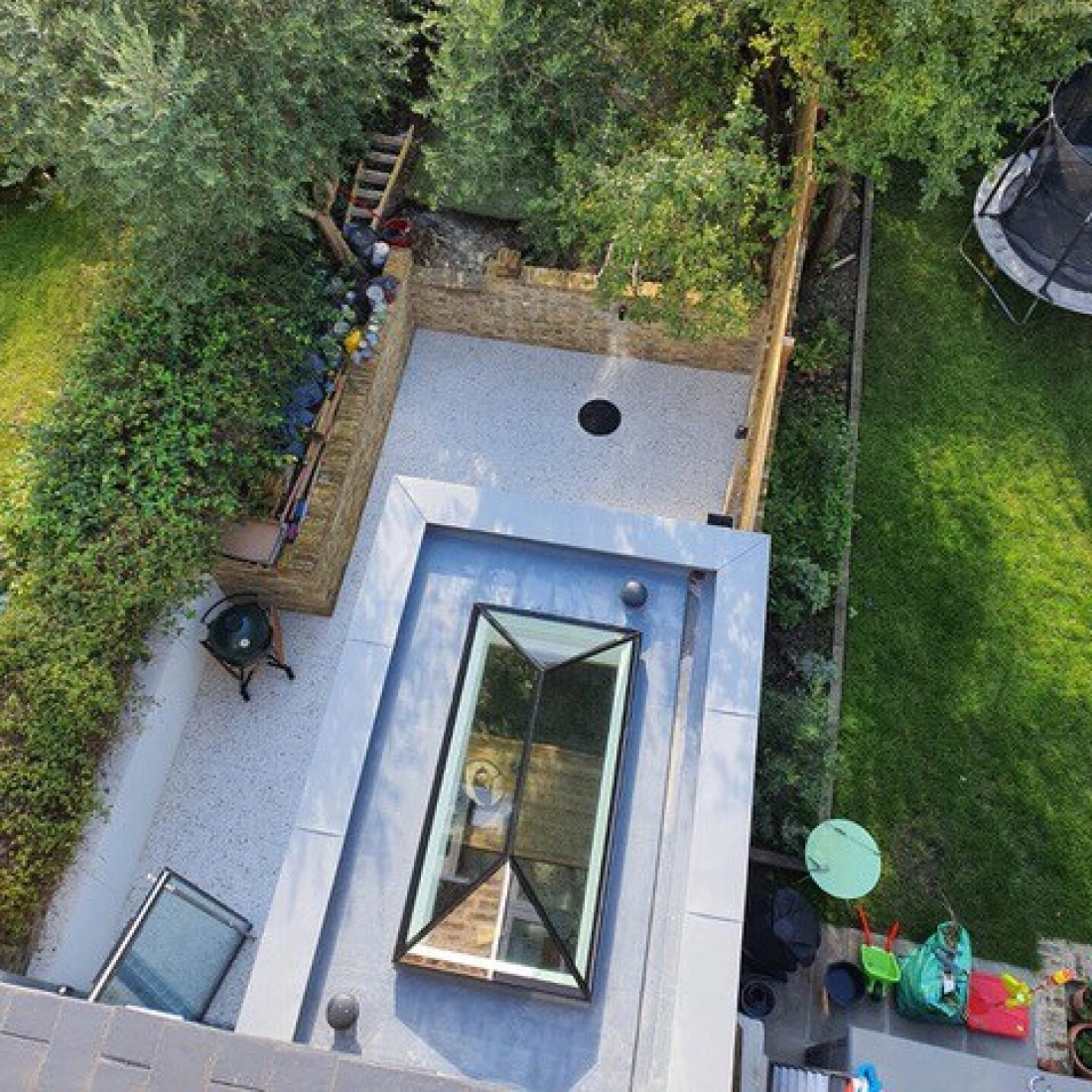 Vista aérea de un jardín con áreas de césped y piedras. Hay un pequeño círculo en el suelo, un patio de grava y un tragaluz en el techo. Al fondo, se ven árboles y un área de juegos.