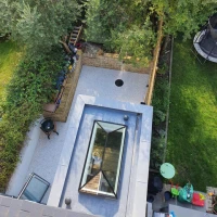 Vue aérienne d'un jardin avec une terrasse en pierre, un trou circulaire au centre, entouré de plantes et d'arbres. Une structure en verre sur le toit permet de voir le jardin. Des meubles de jardin et un trampoline sont visibles.