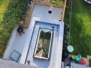 Vista aérea de un jardín con áreas de césped y piedras. Hay un pequeño círculo en el suelo, un patio de grava y un tragaluz en el techo. Al fondo, se ven árboles y un área de juegos.