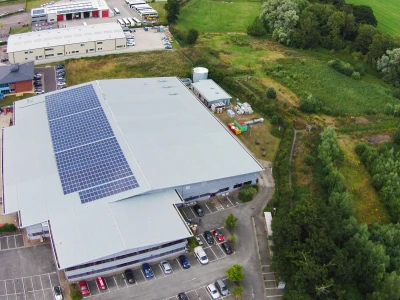 Vista aérea de un edificio comercial con paneles solares en el techo. Se observa un amplio estacionamiento lleno de vehículos y una zona verde con árboles y campo al fondo.