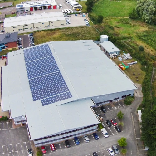 Vista aérea de un edificio comercial con paneles solares en el techo. Se observa un amplio estacionamiento lleno de vehículos y una zona verde con árboles y campo al fondo.