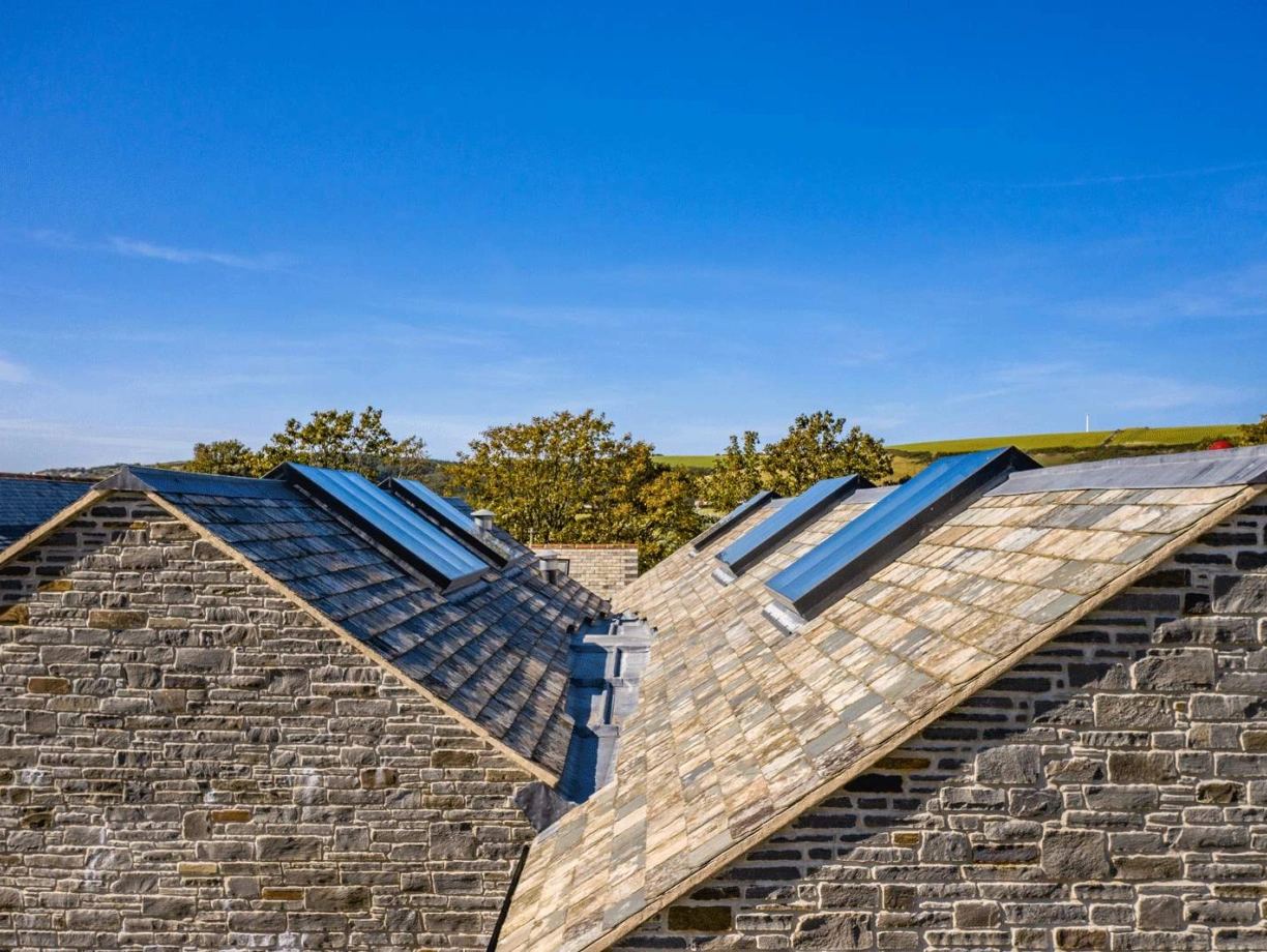 Aerial view of two sloped roofs made of slate tiles, featuring skylights. The backdrop shows a clear blue sky and green hills in the distance.