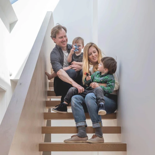 A family of four sits on a staircase. A father and mother hold their young children, enjoying a playful moment. The interior features modern, minimalist design with natural light coming from a skylight above.