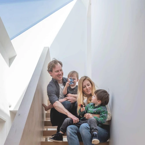 Eine Familie sitzt auf einer Holztreppe. Ein Mann und eine Frau halten jeweils ein Kind in den Armen. Weitere Kinder spielen auf der Treppe. Hinter ihnen ist eine helle, moderne Innenarchitektur mit großen Fenstern.