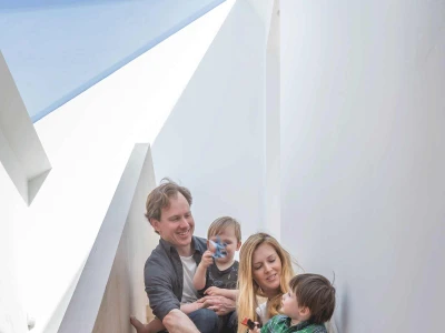 Una familia de cuatro personas sentada en escaleras de madera. Un hombre y una mujer sonríen mientras sostienen a dos niños pequeños. La decoración es moderna y luminosa, con luz natural que entra por una ventana.