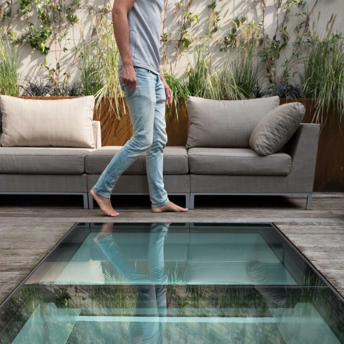 Ein Mann geht barfuß auf einem Glastreppenabsatz, umgeben von einer modernen Terrasse mit gemütlichen Sofas und grünen Pflanzen im Hintergrund.