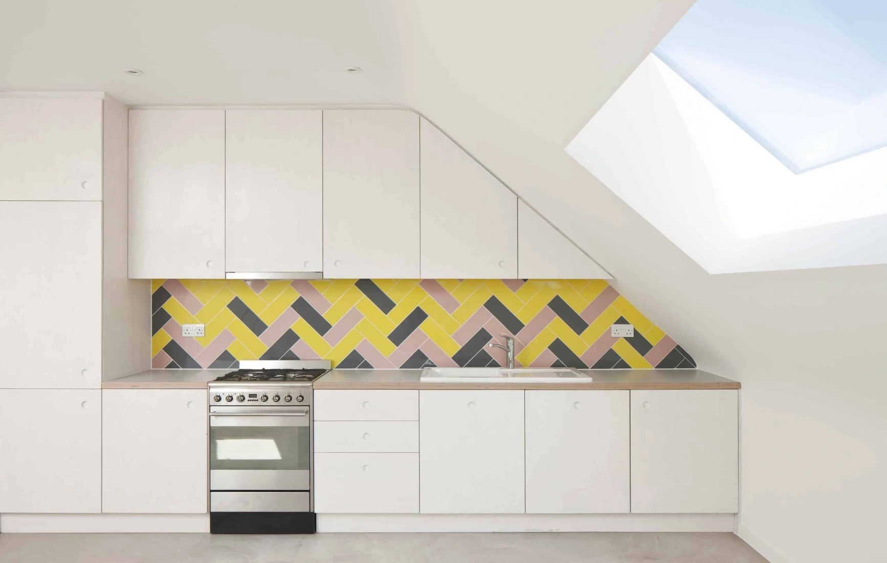 Moderne Küche mit einer schrägen Decke. Weiße Schränke und eine stylische, gelbe Fliesenwand im Fischgrätenmuster. Ein Herd aus Edelstahl steht vor der Fliesenwand, während ein Dachfenster Tageslicht hereinlässt.