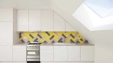 Moderne Küche mit einer schrägen Decke. Weiße Schränke und eine stylische, gelbe Fliesenwand im Fischgrätenmuster. Ein Herd aus Edelstahl steht vor der Fliesenwand, während ein Dachfenster Tageslicht hereinlässt.