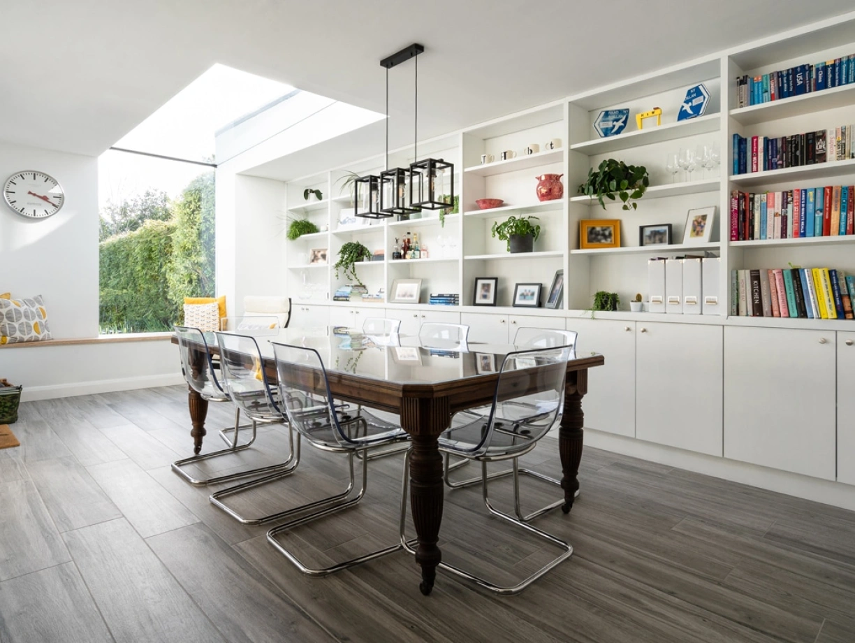A modern dining area featuring a wooden table with transparent chairs. A large window illuminates the space, while shelves filled with books and decorative items line the walls. A cozy seating nook with yellow cushions is visible in the background.