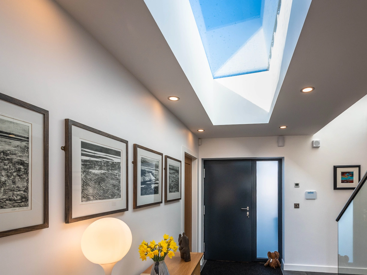 A modern hallway with a sleek design. The space features a large skylight illuminating the area, several framed artworks on the wall, a wooden console table with a white lamp and a vase of yellow flowers, and a dark front door. A small rug and a decorative figure are visible near the entrance.