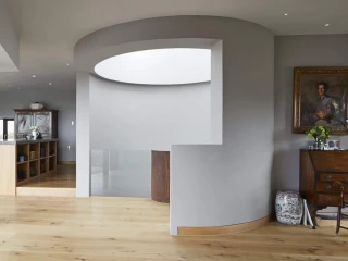 Moderner Wohnraum mit offener Raumgestaltung. Holzboden und große Fenster schaffen eine helle Atmosphäre. Zu sehen sind runde Wände, Säulen und eine Kombination aus modernen und klassischen Möbeln. Ein Essbereich ist im Hintergrund sichtbar.