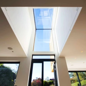 A modern skylight creating a rectangular opening in a white ceiling, allowing natural light to flood in. The view outside shows clear blue skies and greenery. Large windows frame the scene below, enhancing the airy feel of the space.