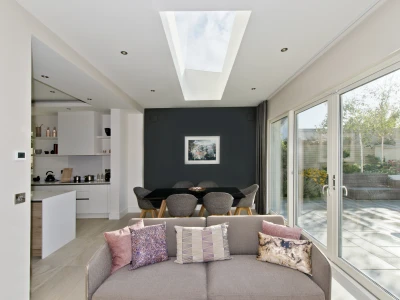 A modern interior showcasing an open living space with a couch adorned with decorative pillows. In the background, a dining area features a black table surrounded by gray chairs, and large glass doors lead to a garden. A skylight above provides natural light, illuminating the room.