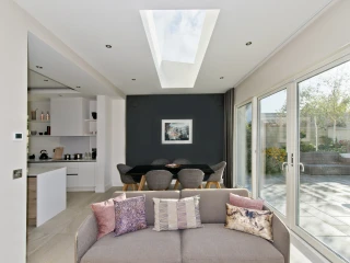 Heller Wohnraum mit einem Sofa und bunten Kissen. Hinter dem Sofa befindet sich ein Essbereich mit einem schwarzen Tisch und grauen Stühlen. Große Fenster und ein Dachfenster sorgen für viel Tageslicht.