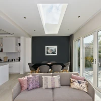 A modern interior showcasing an open living space with a couch adorned with decorative pillows. In the background, a dining area features a black table surrounded by gray chairs, and large glass doors lead to a garden. A skylight above provides natural light, illuminating the room.
