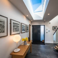 Un hall d’entrée moderne avec des murs blancs. Des œuvres encadrées ornent le mur de gauche. Un éclairage doux provient d’une lampe posée sur un meuble en bois. Une porte noire mène à l’extérieur, et un puits de lumière laisse entrer la lumière naturelle. Des fleurs jaunes dans un vase ajoutent une touche de couleur.