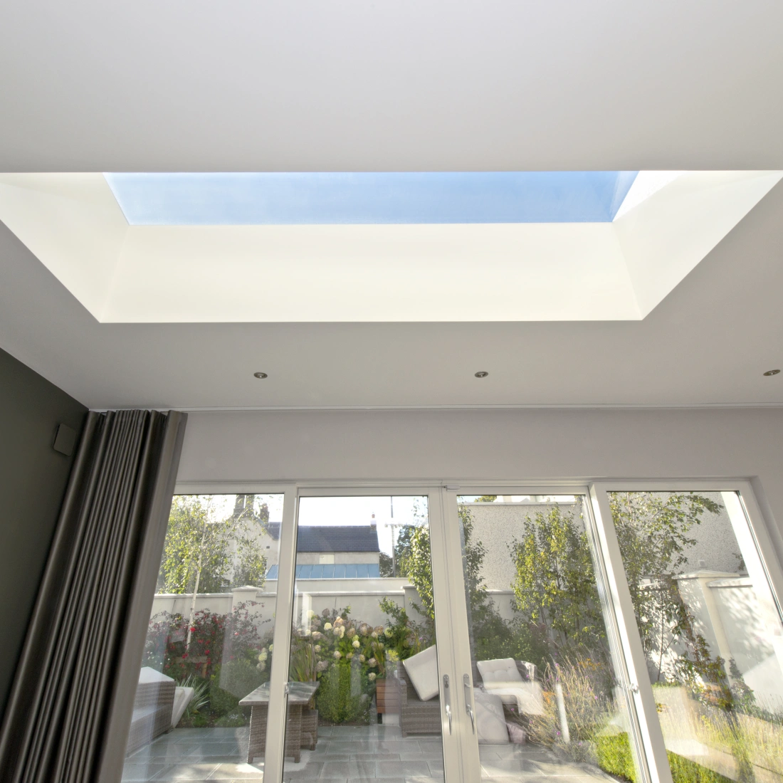Habitación luminosa con un tragaluz rectangular en el techo. Ventanas de vidrio en la pared opuesta, mostrando un jardín exterior con plantas y muebles. Paredes en tonos claros y oscuros.