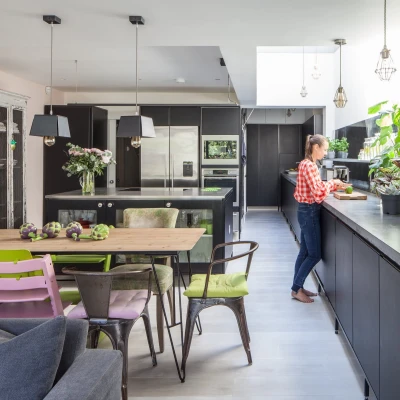 A modern kitchen featuring black cabinets and a large skylight. A woman in a checkered shirt stands at a sleek counter, preparing food. The dining area includes a rustic table surrounded by colorful chairs, with plants and flowers adding decor throughout the space.