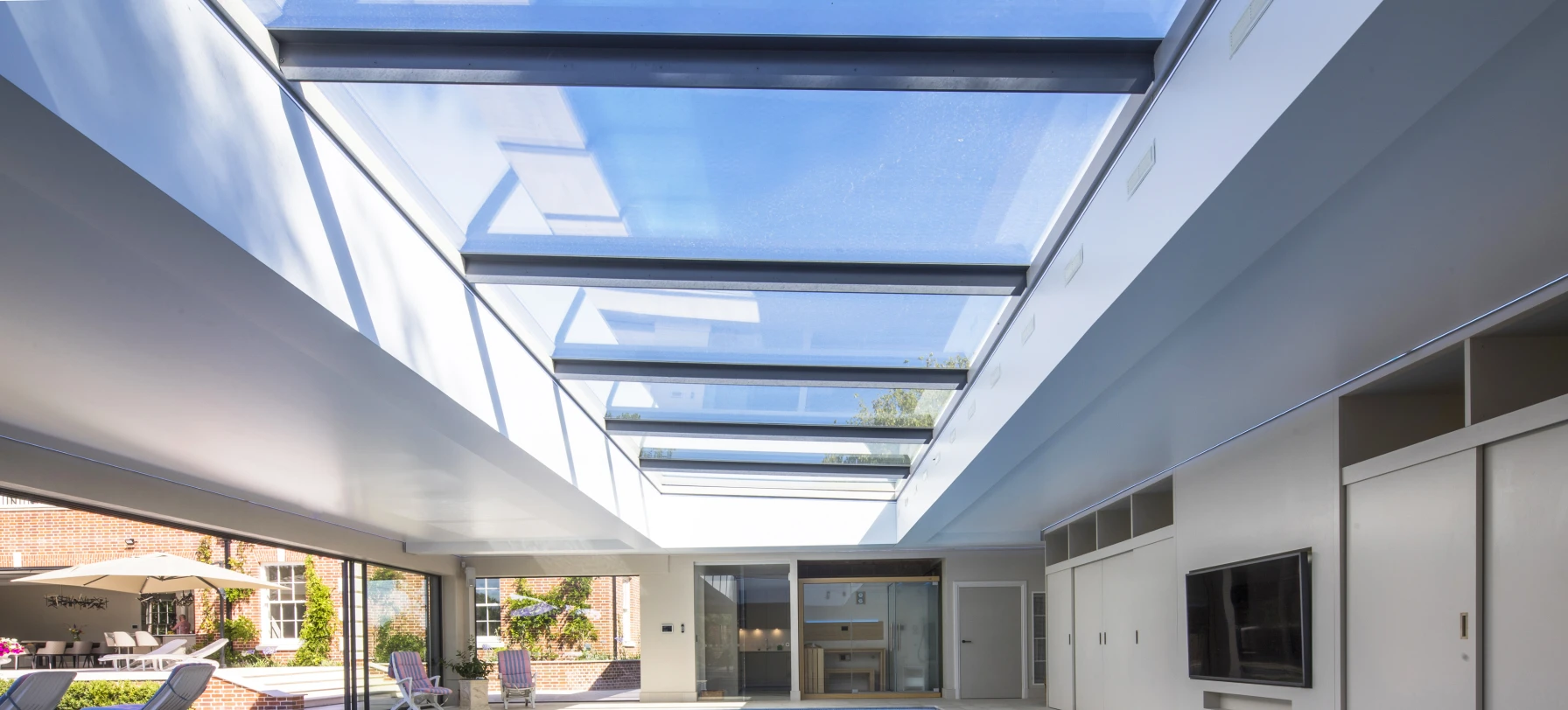Interior de una casa con piscina iluminada por un techo de vidrio. Las paredes son de color claro y hay un área de descanso con sillas. Se puede ver el exterior con un jardín al fondo.