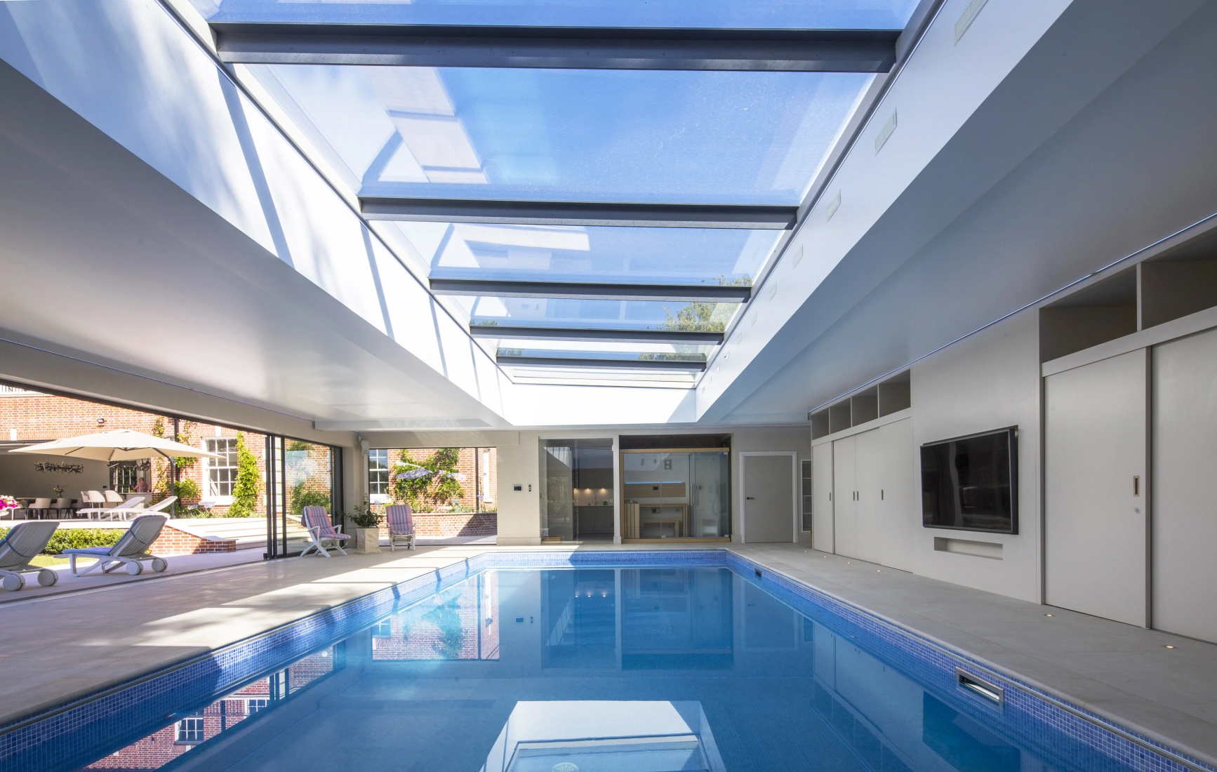 Intérieur d'une piscine moderne avec un plafond vitré, baignée de lumière naturelle. Des chaises longues sont disposées sur le côté et des portes coulissantes mènent à l'extérieur.