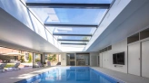 Interior de una casa con piscina iluminada por un techo de vidrio. Las paredes son de color claro y hay un área de descanso con sillas. Se puede ver el exterior con un jardín al fondo.