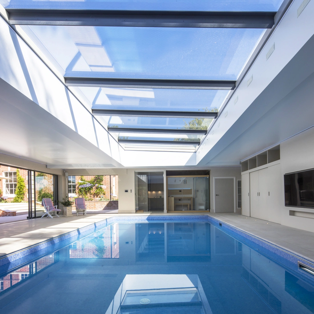 Intérieur d'une piscine moderne avec un plafond vitré, baignée de lumière naturelle. Des chaises longues sont disposées sur le côté et des portes coulissantes mènent à l'extérieur.