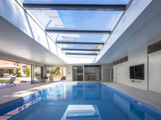 Innenansicht eines modernen, hellen Schwimmbades mit großen Fenstern und einem Glasdach. Das Wasser reflektiert das Licht. Es gibt Liegen am Rand des Beckens und einen Fernseher an der Wand. Die Umgebung wirkt elegant und einladend.