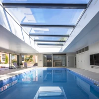 A spacious indoor pool area with a bright skylight, featuring a clear blue pool and modern minimalist design. Sliding doors lead to an outdoor space with lounge chairs.