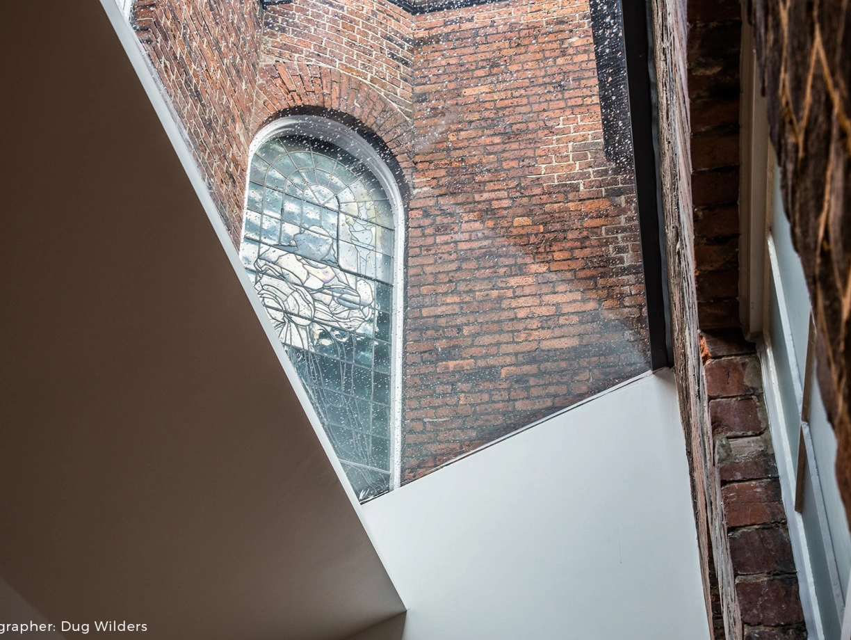 Ein Blick auf ein historisches, mit Mauerwerk verziertes Gebäude. Ein Fenster aus buntem Glas ist sichtbar, während Licht durch einen modernen Dachaufbau strömt.