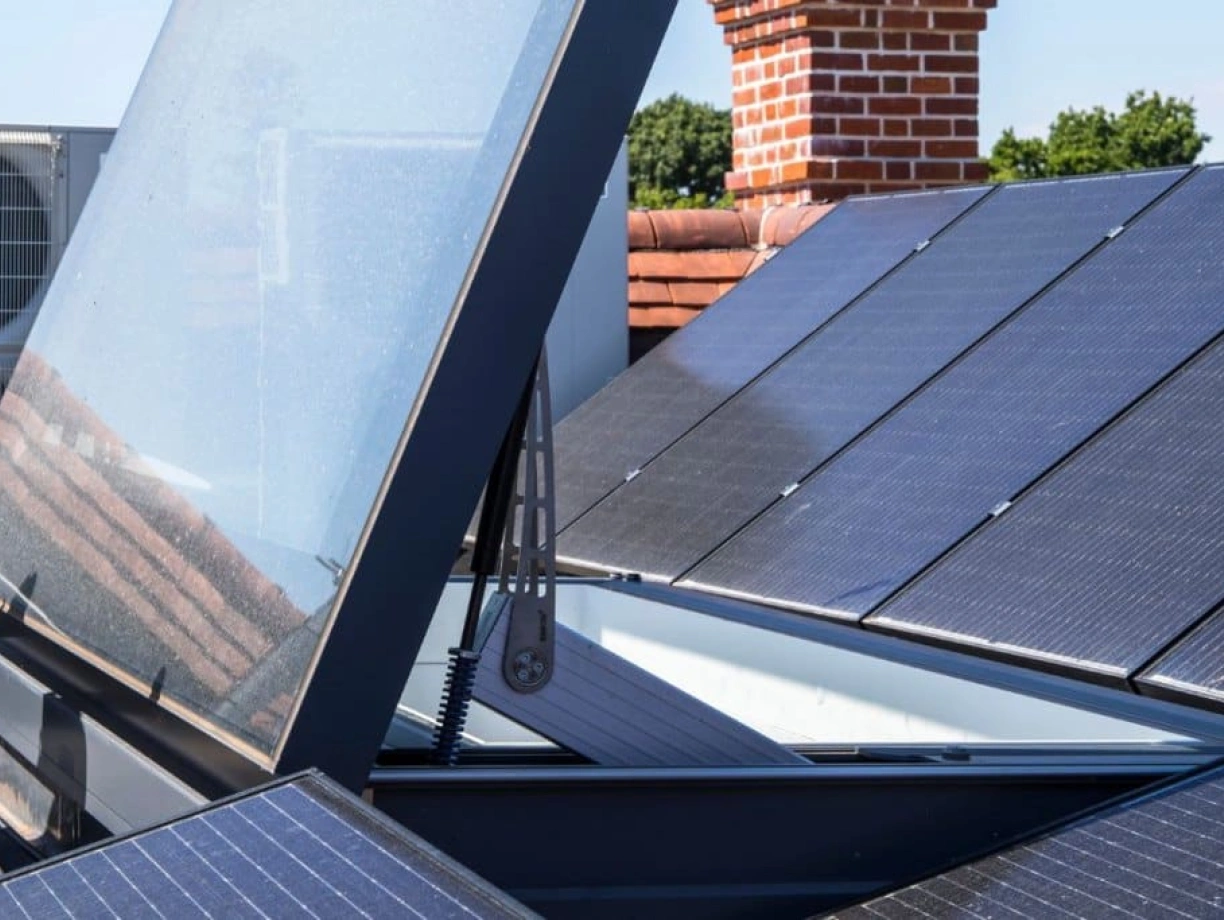 Solar panels installed on a rooftop, with one panel tilted open to expose a skylight or vent below. A red brick chimney is visible in the background, along with an air conditioning unit.
