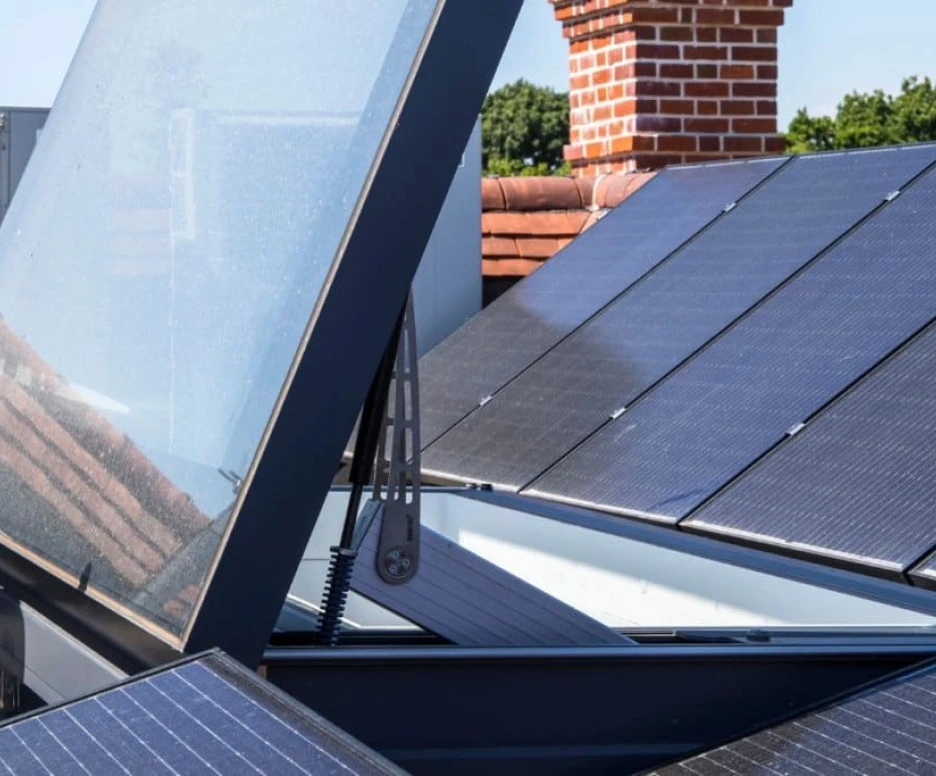 Paneles solares en un techo, con una estructura que parece abrirse y un fondo de cielo azul. Se observa un sistema de refrigeración al fondo y ladrillos rojos en el edificio.