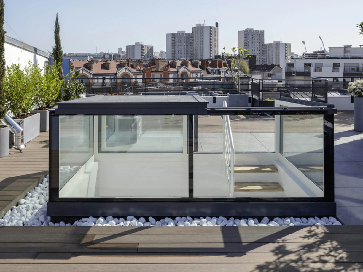 A modern rooftop terrace with a glass structure, surrounded by greenery and decorative white stones. In the background, city buildings and blue sky are visible.