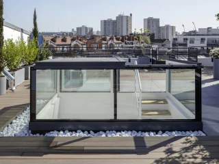 A modern rooftop terrace featuring a glass-walled structure surrounded by greenery and white stones. The skyline shows residential buildings in the background under a clear blue sky.