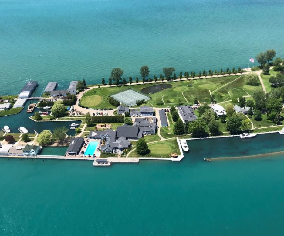 Aerial view of a lush, green island with waterfront properties, boats docked along the shore, and a swimming pool visible. The vibrant turquoise water surrounds the island, which features trees and neatly landscaped lawns.