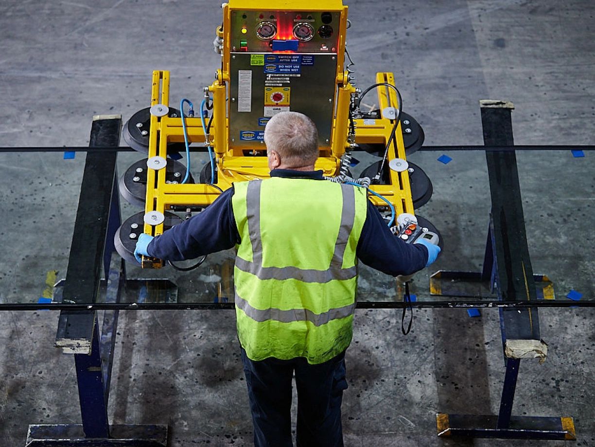 A worker in a reflective vest operates a glass handling machine in a warehouse. The machine is positioned over large sheets of glass, showing various controls and indicators.