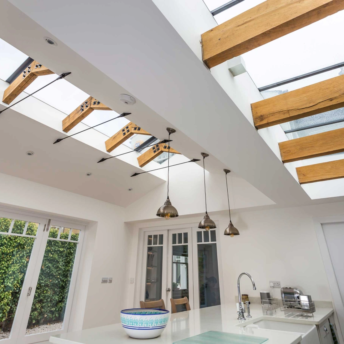 Luminoso cucina con soffitto alto, dotato di finestre a tetto in vetro e travi in legno. Eleganti lampade pendenti d'oro illuminano il piano di lavoro chiaro. Una grande finestra a vetro si affaccia su un giardino verde.