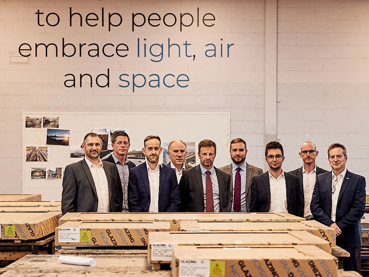 A group of nine professionals stands in a warehouse setting, with wooden crates in the foreground and a motivational wall quote behind them. They are dressed in business attire and appear to be engaged in conversation or discussion.