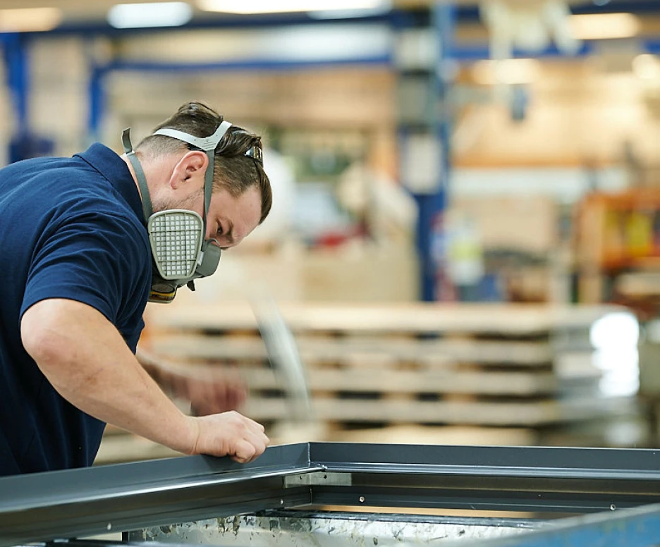 A man wearing a mask and ear protection focuses on assembling a metal frame in a workshop. The background shows shelves filled with materials and tools, indicating a busy manufacturing environment.
